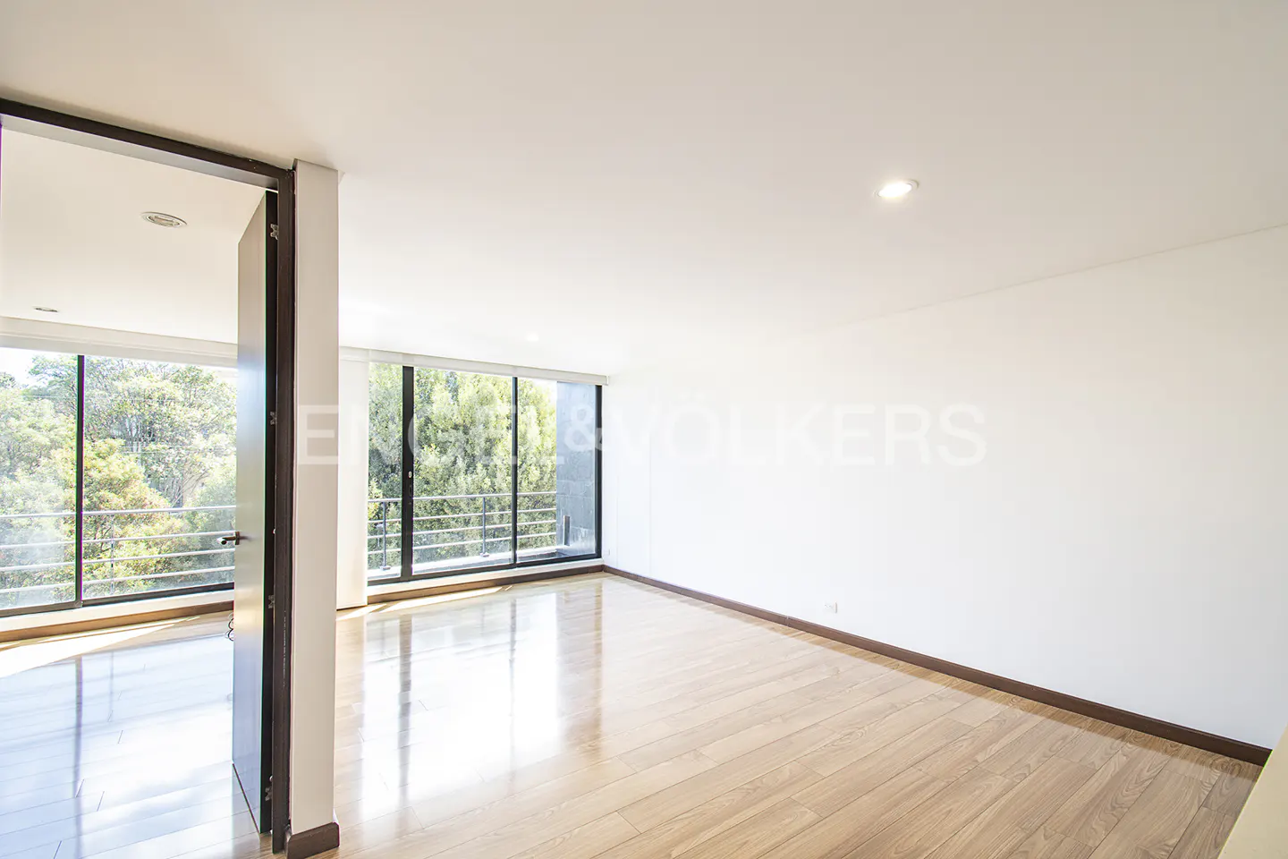 Bright, empty room with light wood floors, white walls, and large windows overlooking trees. Doorway on the left.