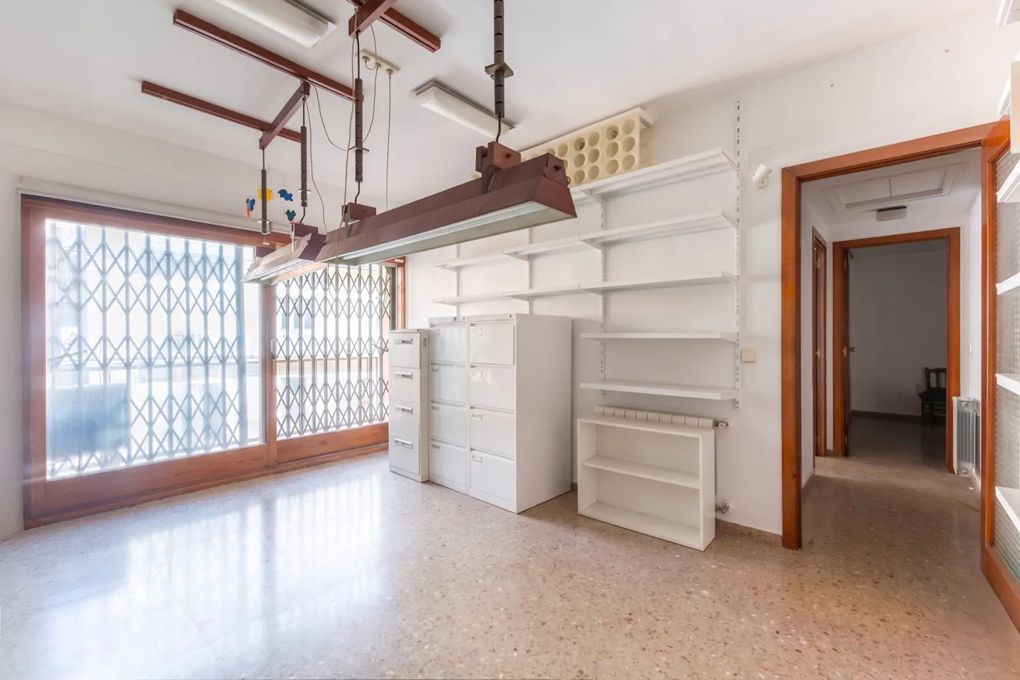 Empty room with white walls, terrazzo floor, and metal security gate on a large window. White shelves and cabinets line the walls.