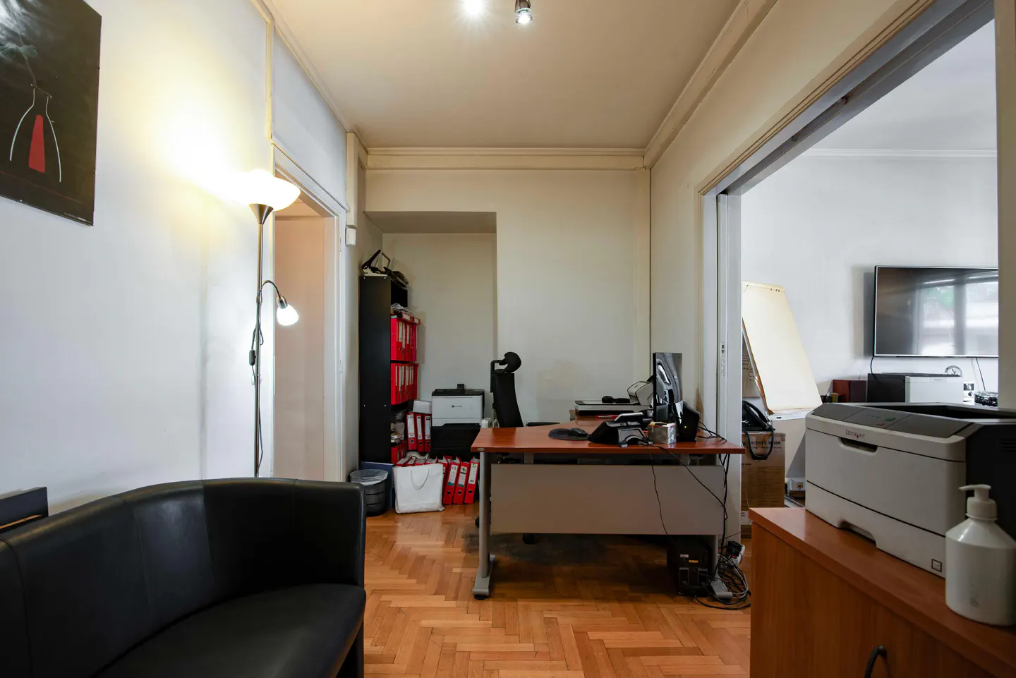 A bright office space with a desk, computer, black sofa, and herringbone wood floors. A printer sits on a wooden cabinet.