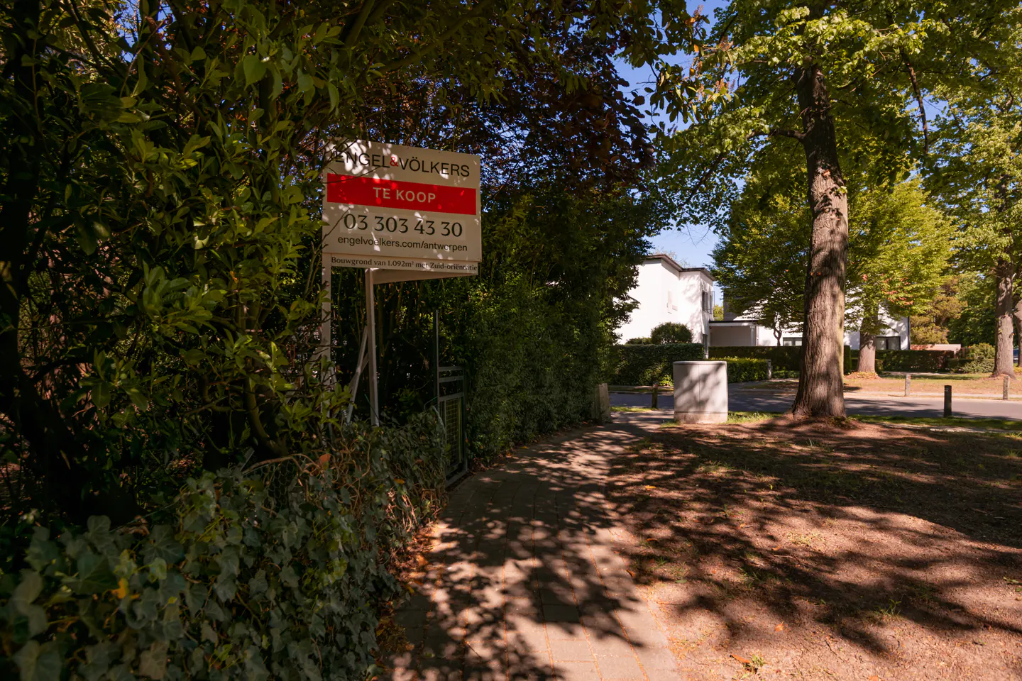 A "Te Koop" (For Sale) sign is posted in front of a property entrance with trees and a path.
