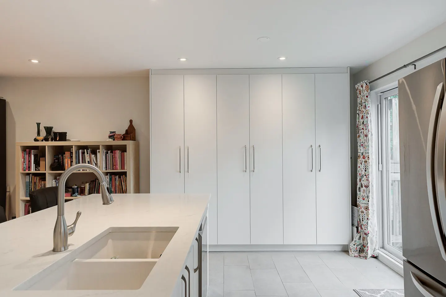 Bright kitchen with white cabinets, marble countertop island with sink and faucet, and stainless steel refrigerator.