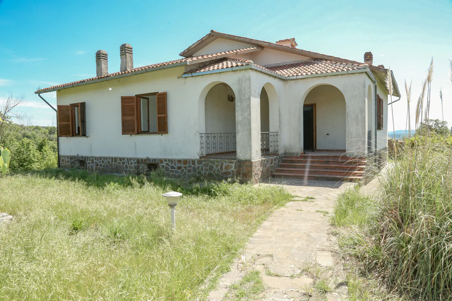 Exterior view of a white, single-story house with a red tile roof and arched porch. The house has brown shutters and a stone foundation. Overgrown grass surrounds the house.