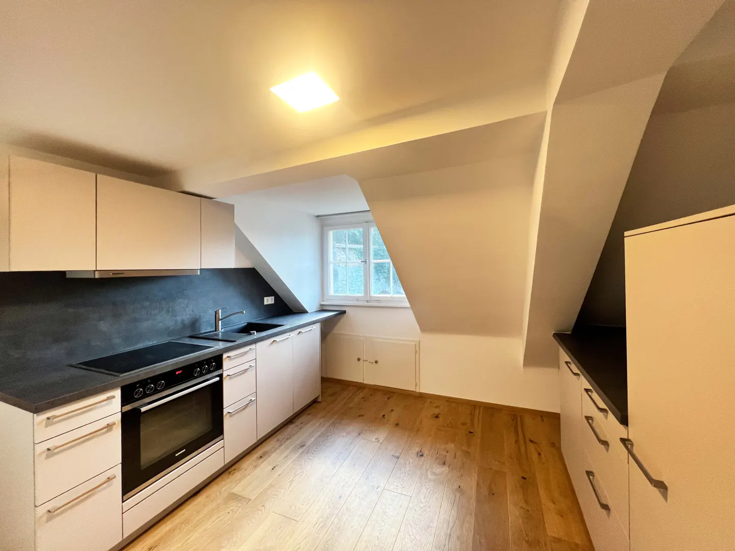 A bright kitchen with light wood floors, white cabinets, and a black countertop. A window is visible in the background.