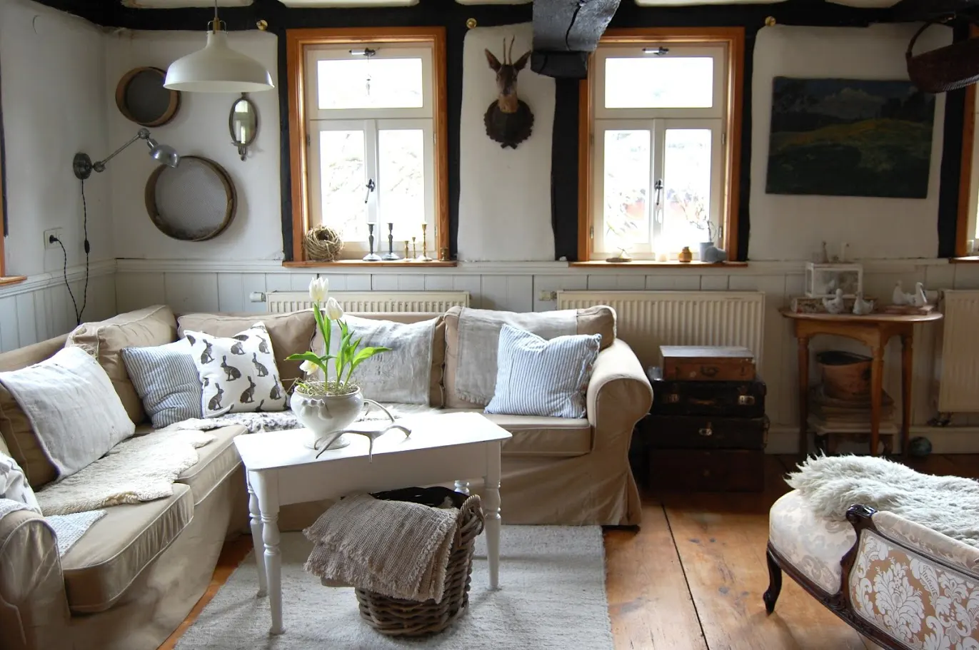 Cozy living room with beige sofas, white walls, and wooden floors. A white coffee table holds flowers, and windows offer natural light.