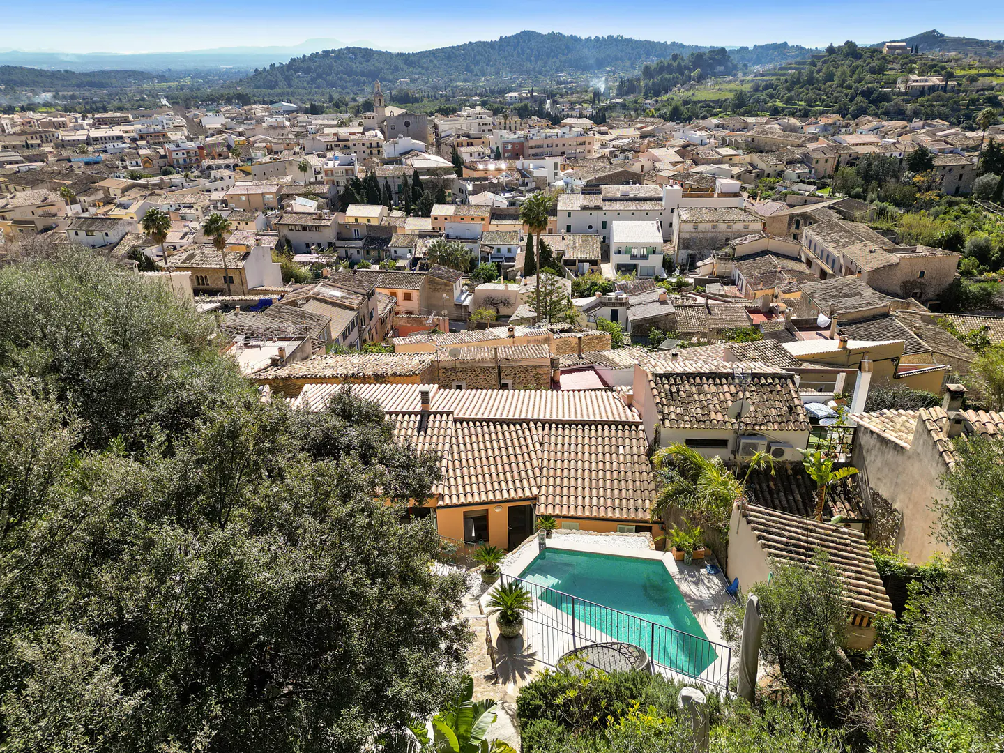 Aerial view of a European town with terracotta rooftops, green hills in the background, and a turquoise pool in the foreground.