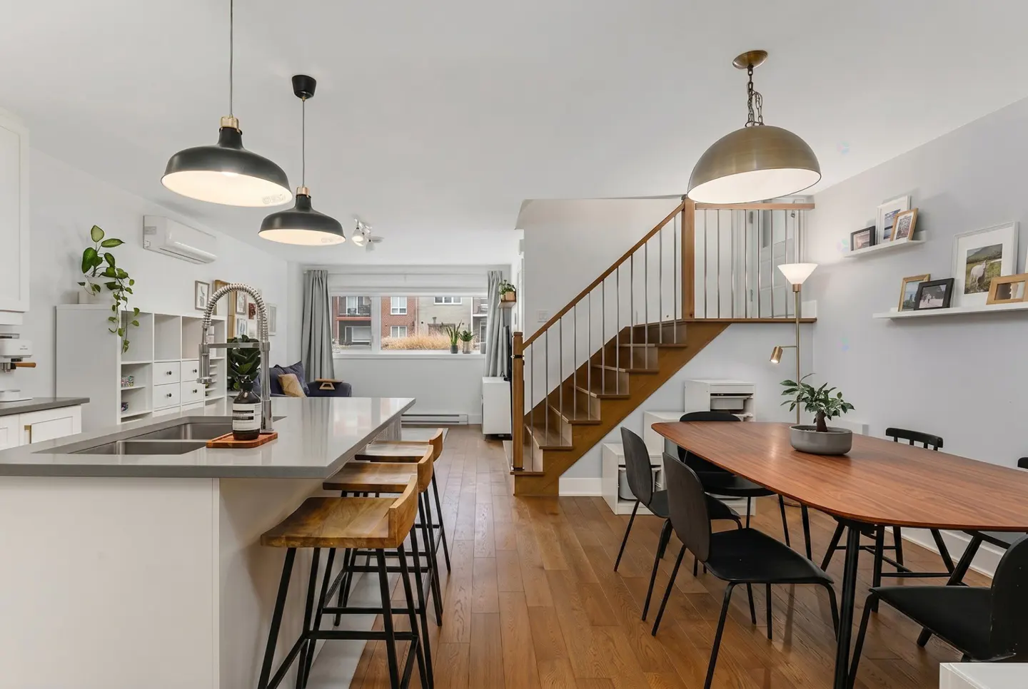 Open-concept living space with a kitchen island, wooden stools, dining table with black chairs, and a staircase. Hardwood floors.