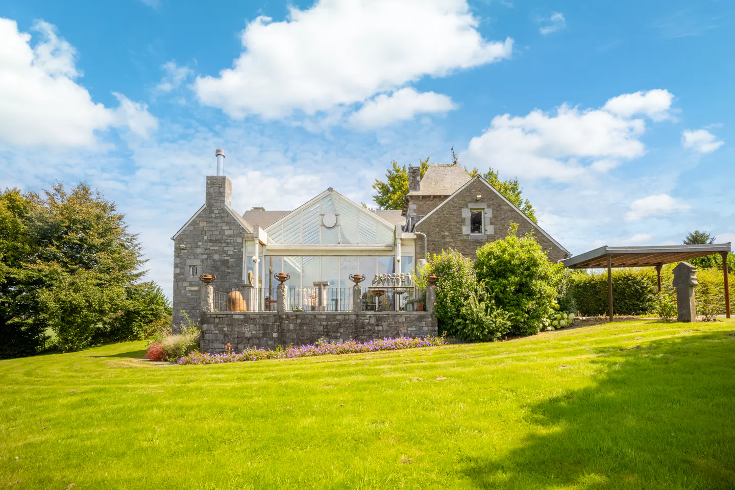 Exterior view of a stone house with a glass conservatory, green lawn, and blue sky with clouds.