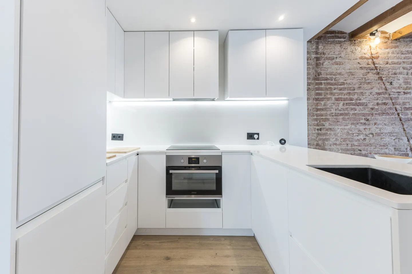 Bright, modern kitchen with white cabinets, countertops, and stainless steel oven. Exposed brick wall with light fixture adds rustic charm.