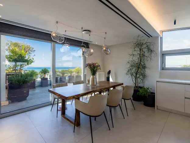Bright dining room with a wood table, beige chairs, and glass pendant lights. A large window shows a patio with ocean views.