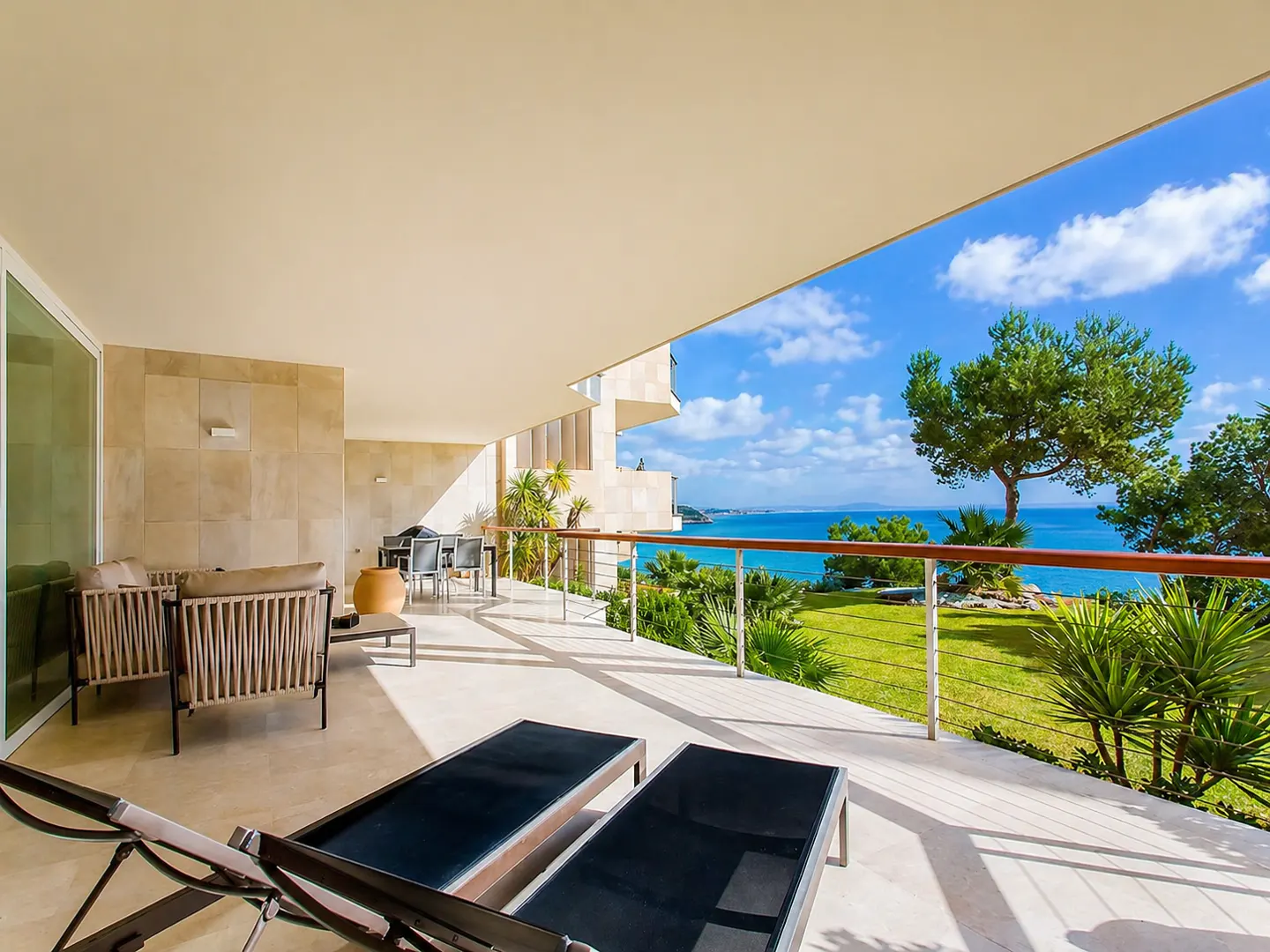 Luxury patio with lounge chairs, sofa, and dining set overlooking the ocean. Blue sky and green lawn visible.