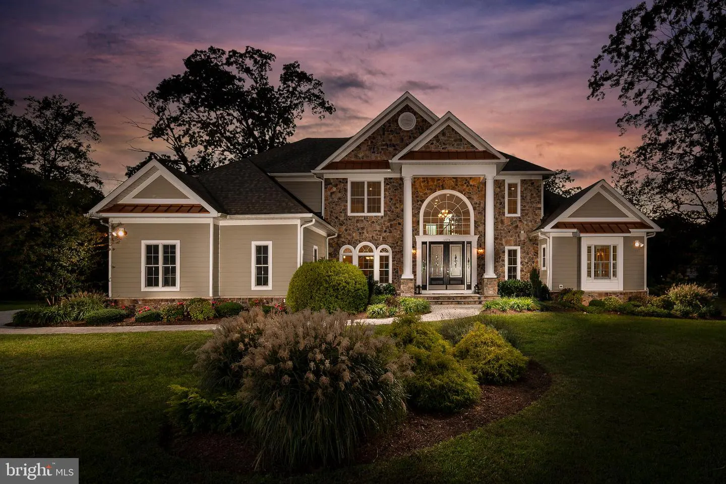 Two-story house with stone and tan siding, white trim, and a dark roof at dusk. A manicured lawn and shrubs are in the front.