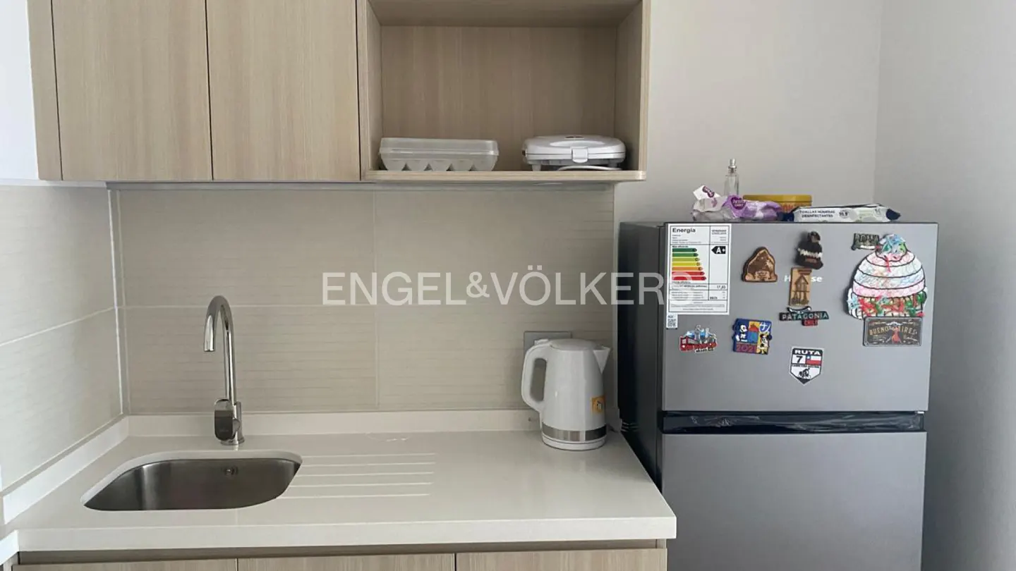A compact kitchen with light wood cabinets, a stainless steel sink, and a silver refrigerator covered in magnets.