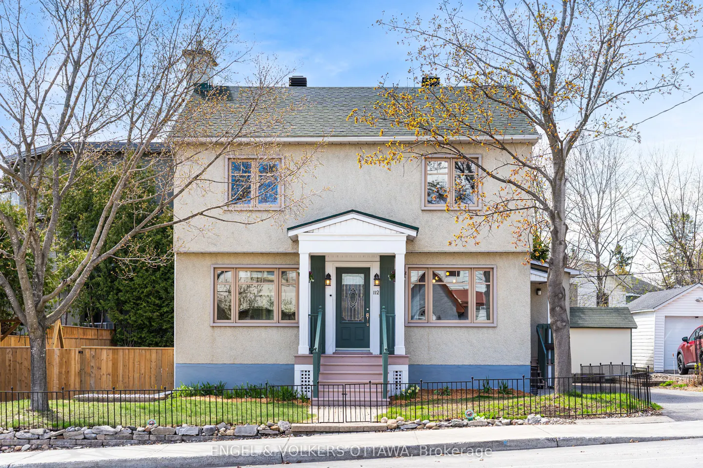 Two-story beige house with a green door and a gray roof, framed by trees under a blue sky.