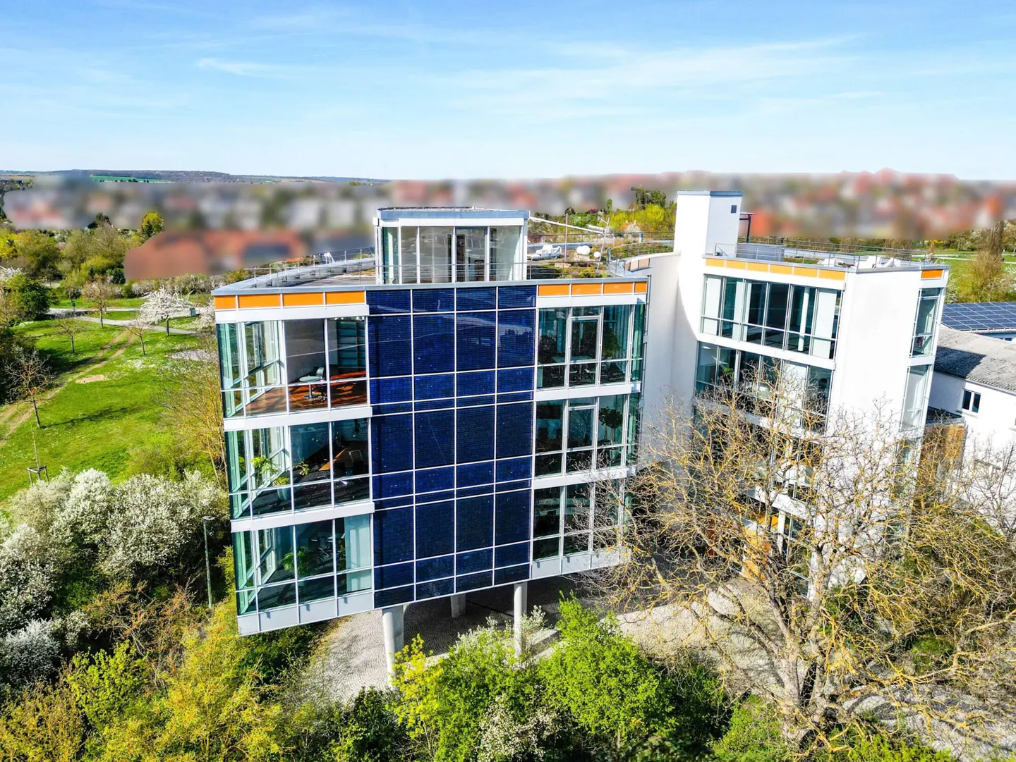 Aerial view of a modern multi-story office building with a blue glass facade, white walls, and surrounding green trees.