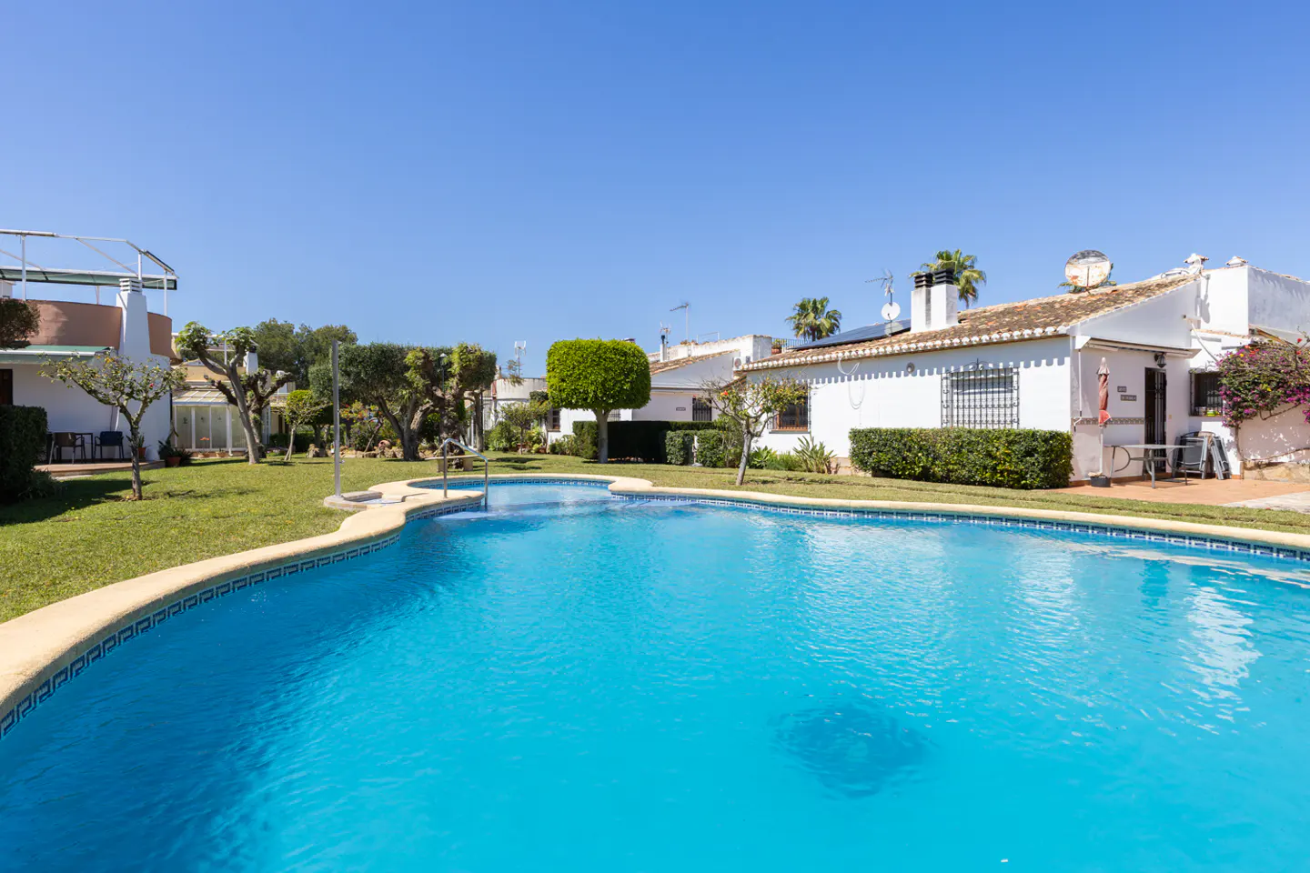 A bright blue swimming pool is surrounded by green grass and white buildings under a clear blue sky.