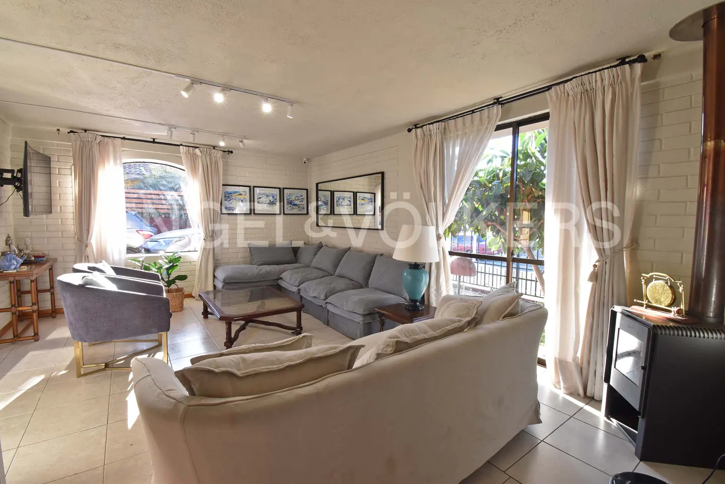 A bright living room with a gray sectional sofa, beige armchairs, and a wood-burning stove. Cream-colored curtains frame the windows.