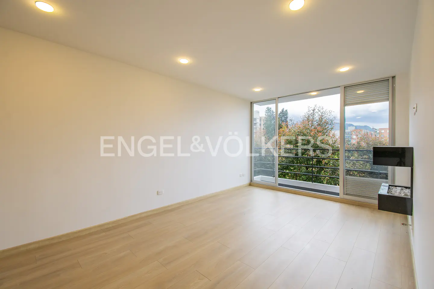 Bright, empty room with light wood floors, white walls, and large windows overlooking trees and a city. Engel & Völkers logo on the wall.