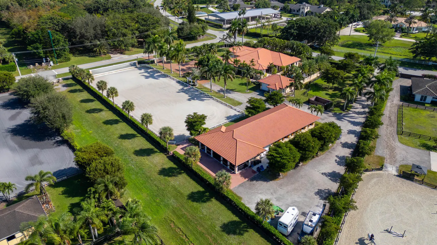 Aerial view of an equestrian estate with a large sand arena, stables, and lush green landscaping. Orange tile roofs top the buildings.