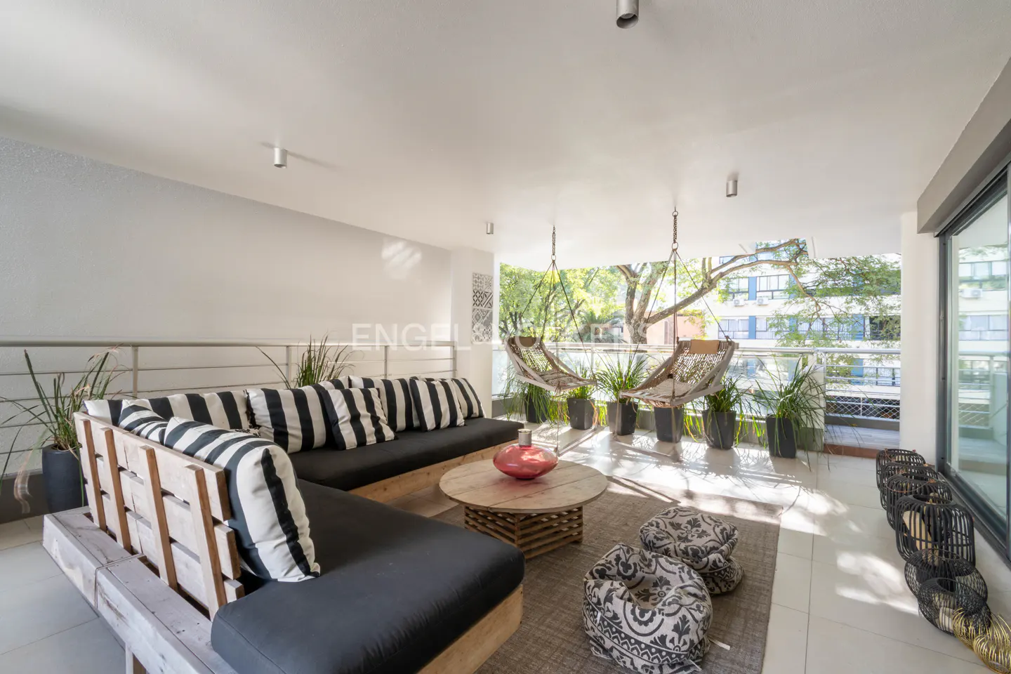 Bright, modern balcony with a wooden couch, black and white striped pillows, and hanging chairs overlooking trees.