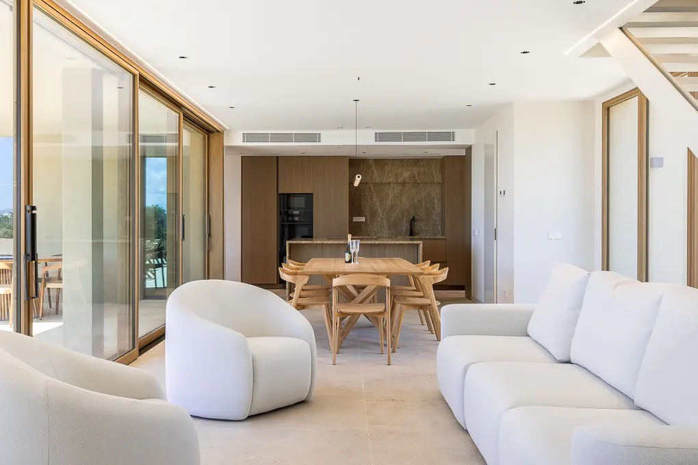 Bright, modern living space with white sofas, wood dining set, and open kitchen. Sliding glass doors lead to a patio.