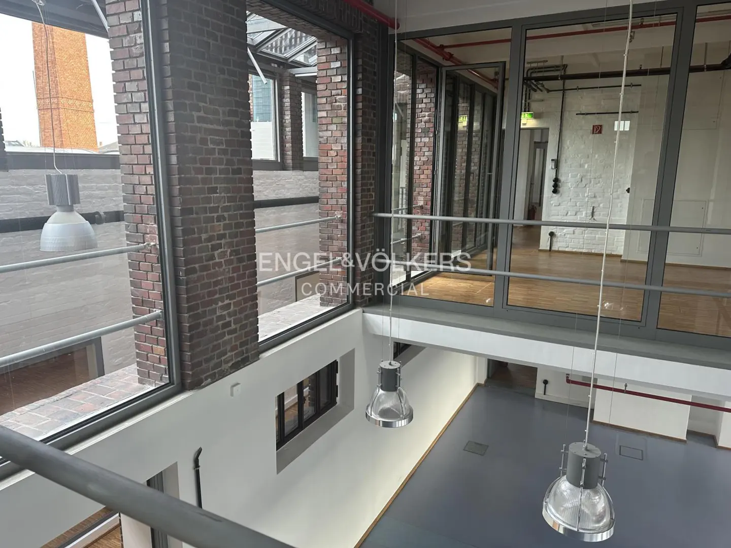 Interior view of a commercial space with brick walls, large windows, and hanging industrial-style lights. The floor is gray, and the space is open and airy.
