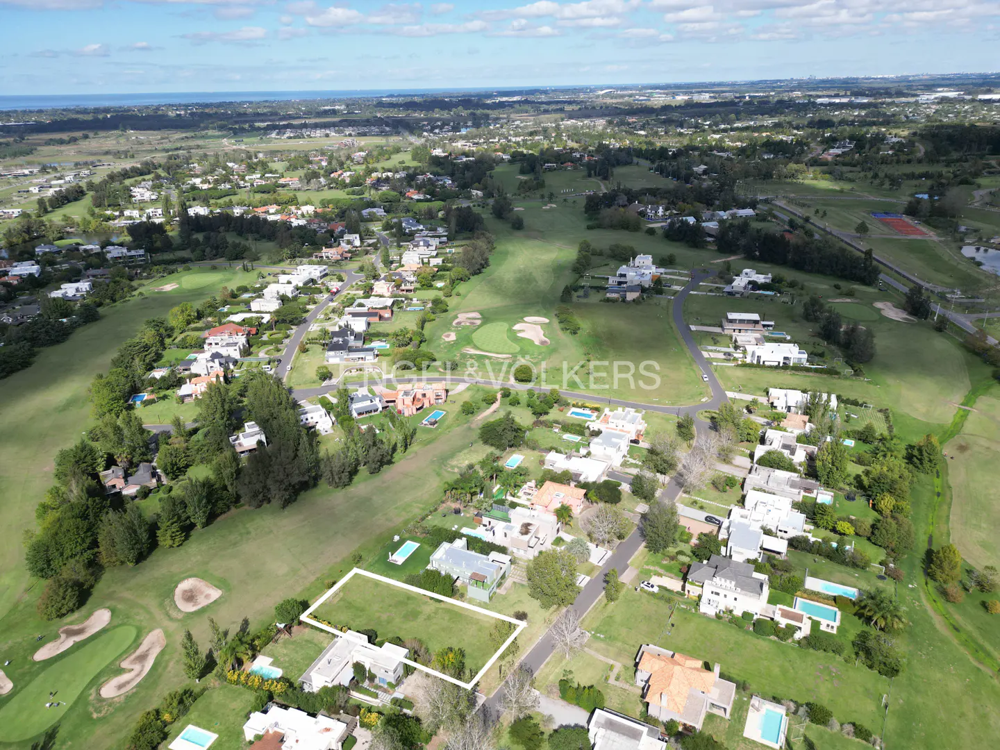 Aerial view of a green residential area with houses, trees, and a golf course. A white rectangle highlights a plot of land.