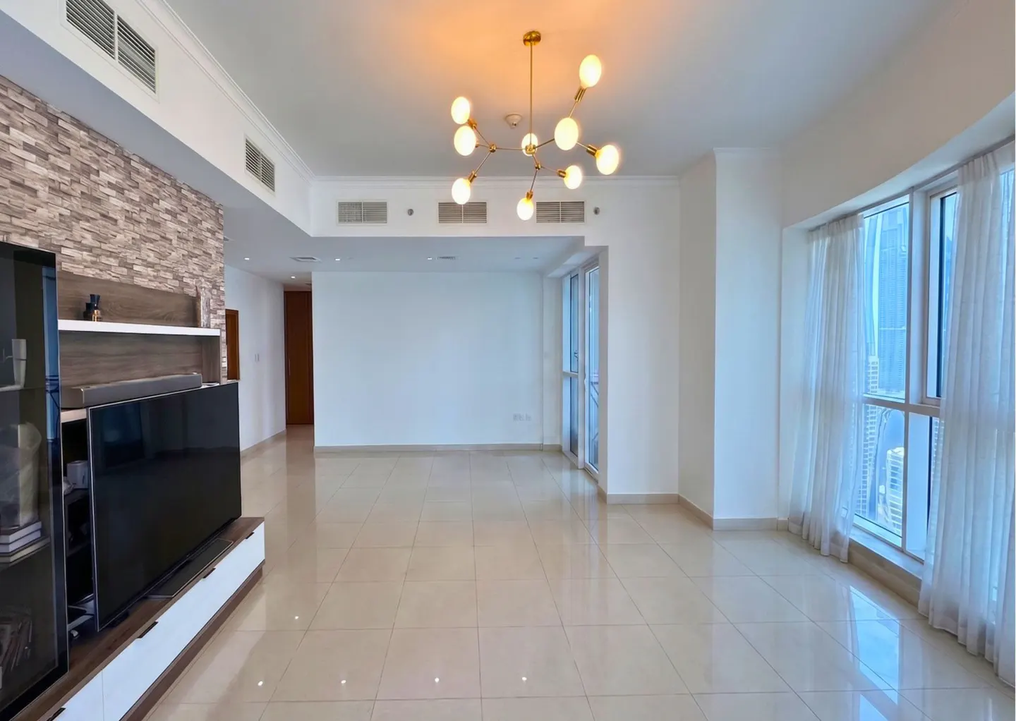 Bright, modern living room with white tile floors, a stone accent wall, a large TV, and a contemporary gold chandelier.