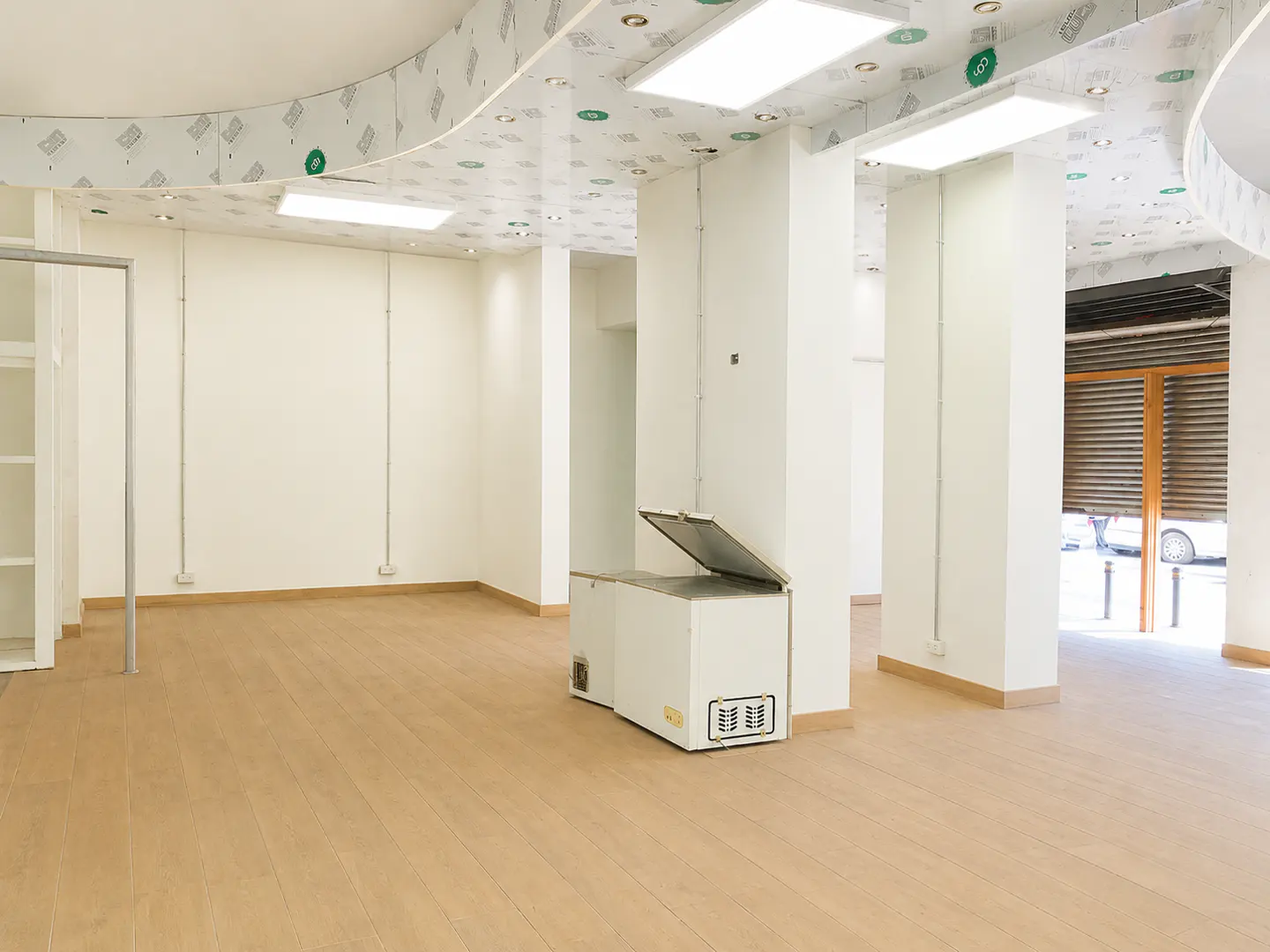 Empty retail space with white walls, light wood floors, a chest freezer, and a curved ceiling with recessed lighting.