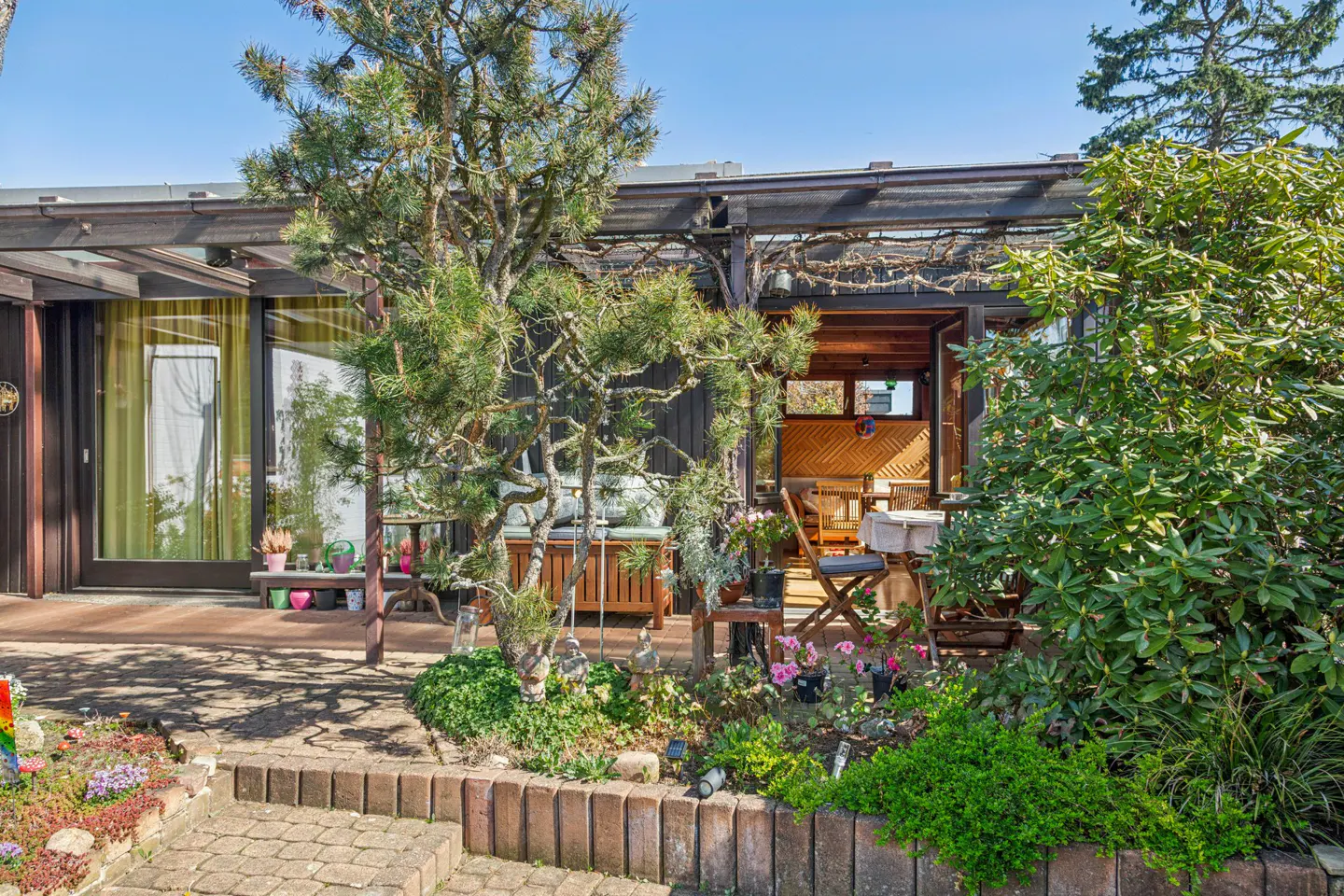 Exterior view of a brown house with a patio, wooden furniture, and lush green plants. Brick steps lead to the patio.