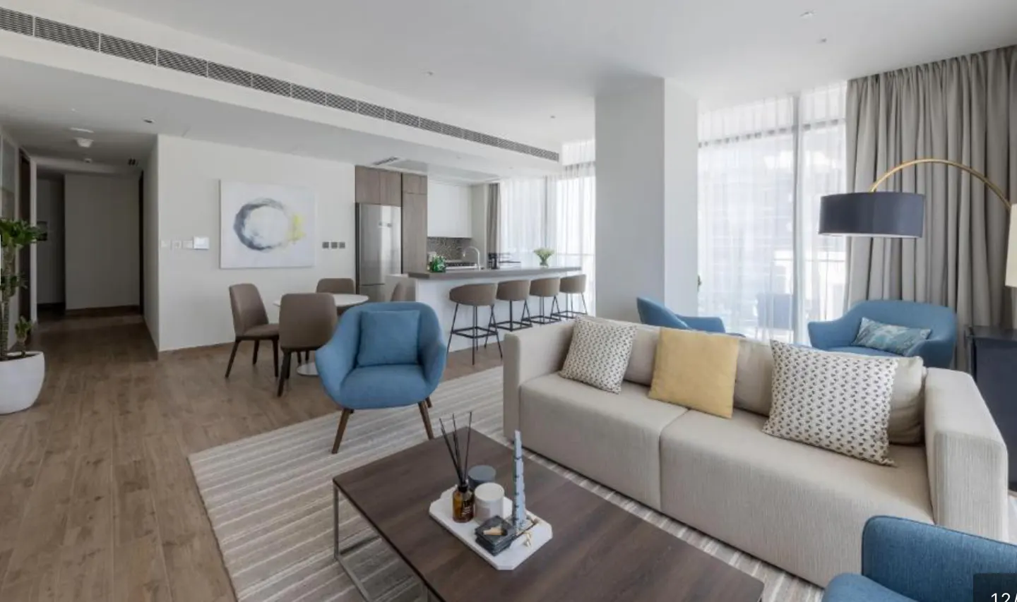 Bright, modern apartment interior with a beige sofa, blue chairs, and a dark wood coffee table. The kitchen has a breakfast bar with stools. Large windows provide natural light.