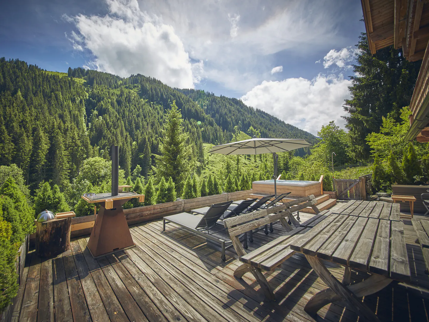 Wooden deck with lounge chairs, table, grill, and hot tub under an umbrella, with a forest and cloudy sky in the background.