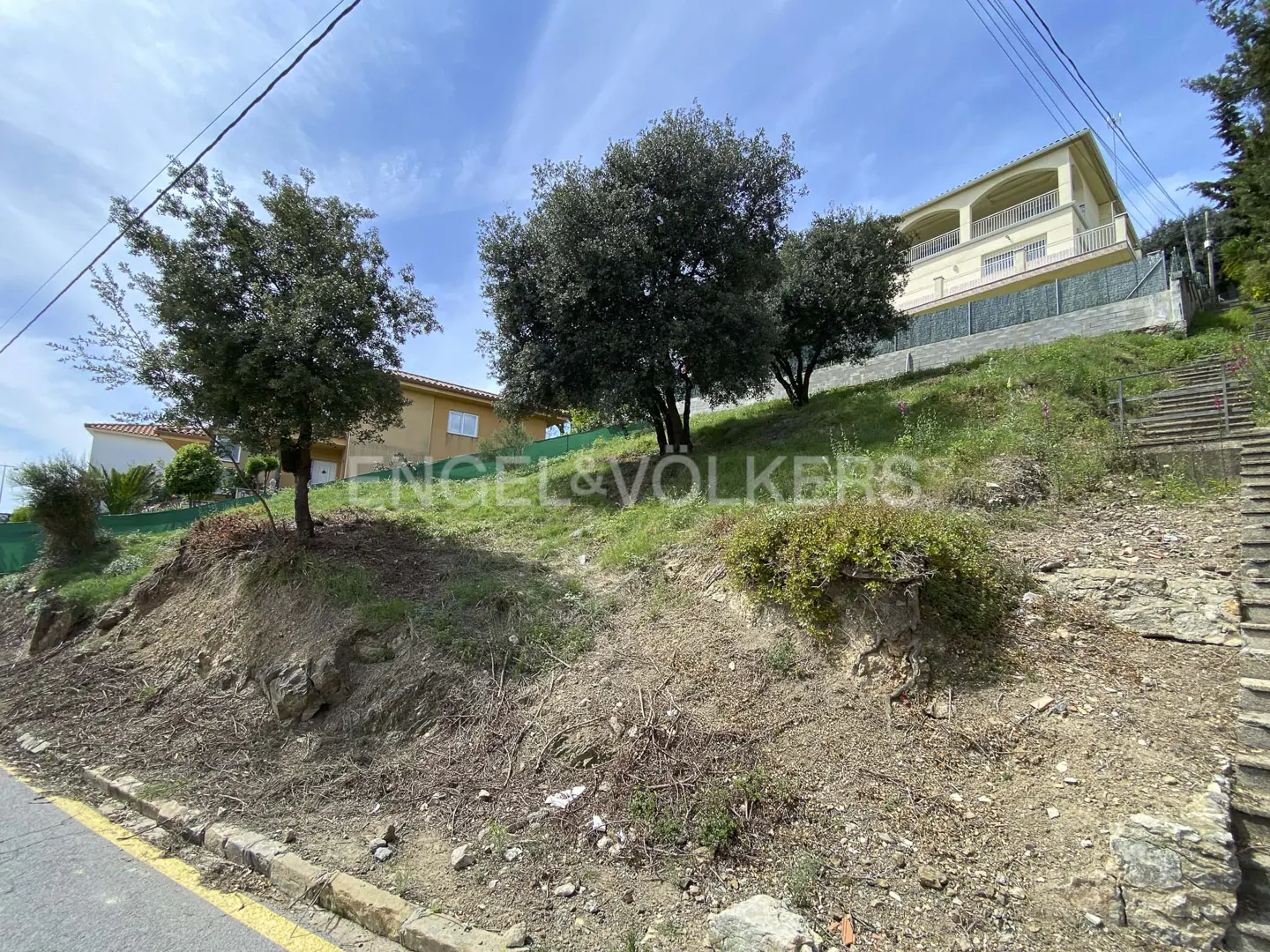 Exterior view of a tan house on a grassy hill with trees under a blue sky. "Engel & Volkers" is printed on the hillside.