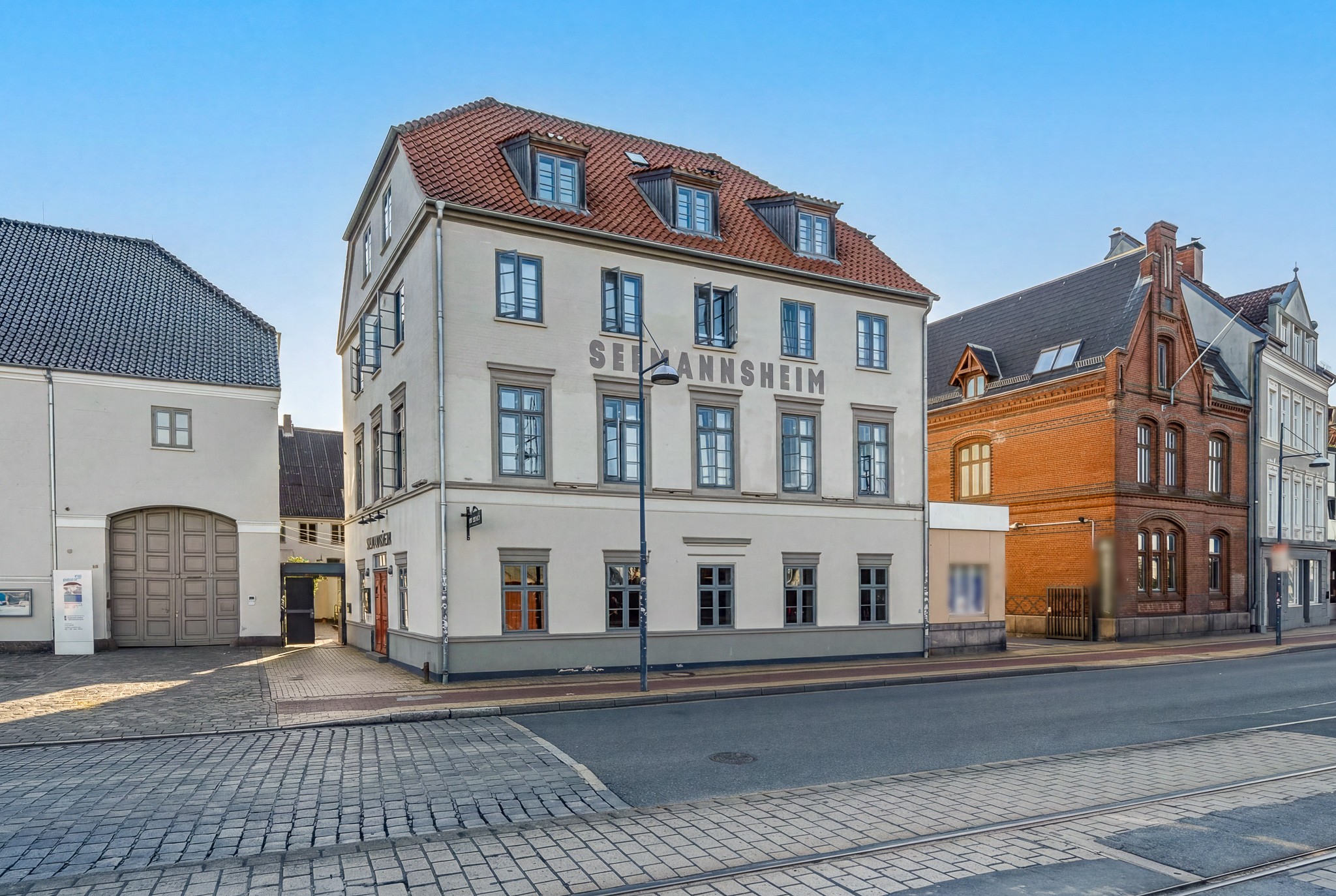 A four-story beige building named "Seimannsheim" with a red tiled roof, situated on a cobblestone street with tram tracks.