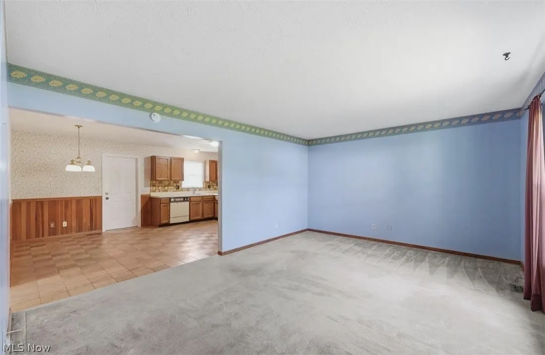 A light blue living room with gray carpet and a leaf-patterned border. An open doorway leads to a kitchen with brown cabinets and a dining area with wood paneling.