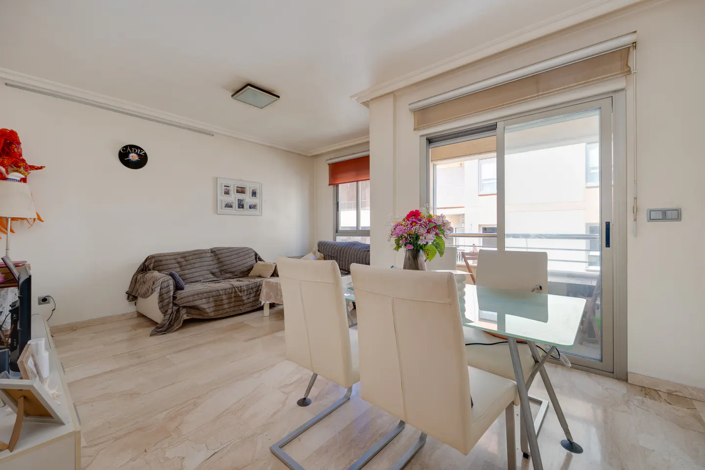 Bright living room with marble floors, a glass table with white chairs, and a brown sofa with a blanket. Sliding glass doors lead to a balcony.