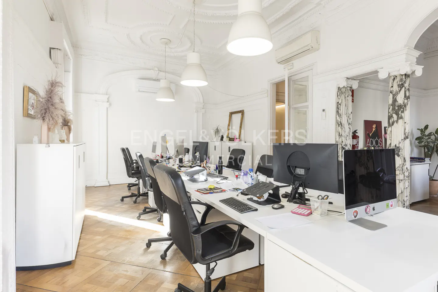 Bright office space with white walls, herringbone wood floors, and rows of desks with computers and black chairs.
