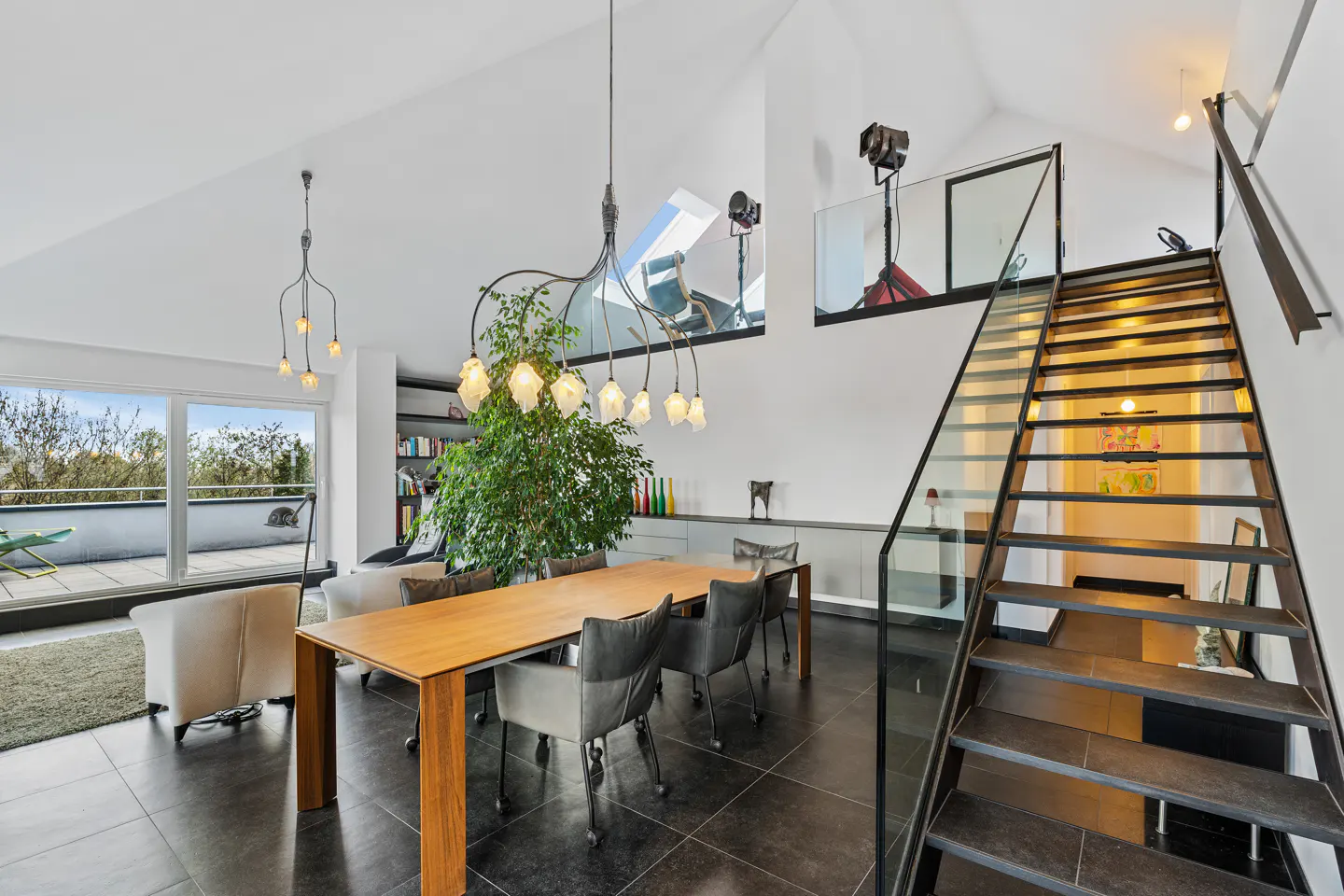Bright, modern dining room with a wood table, gray chairs, and a large green plant. Black tile floors, white walls, and a metal staircase with glass railing.