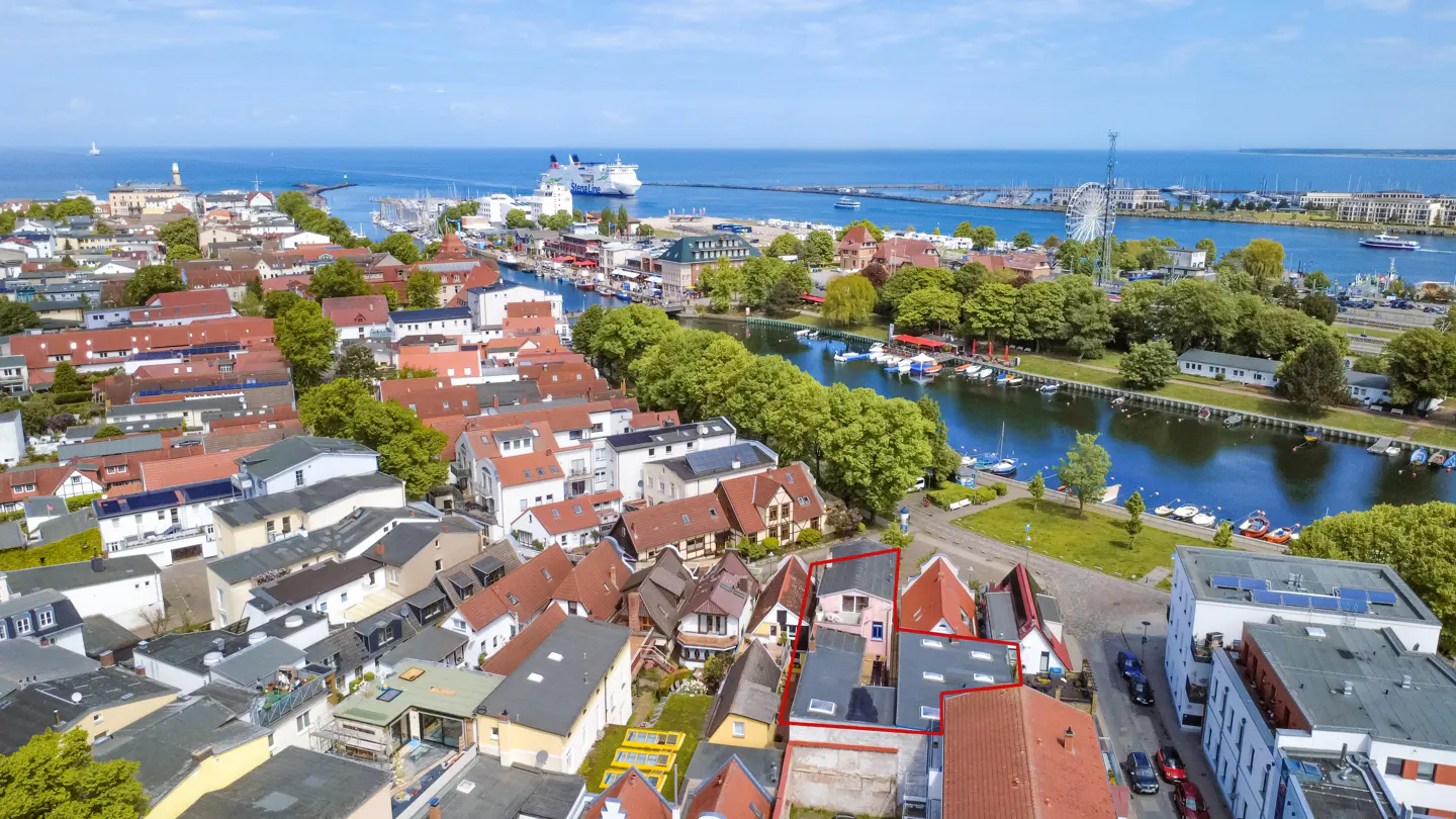 Aerial view of a coastal town with a red-outlined property near a canal, harbor, and ferry under a clear blue sky.