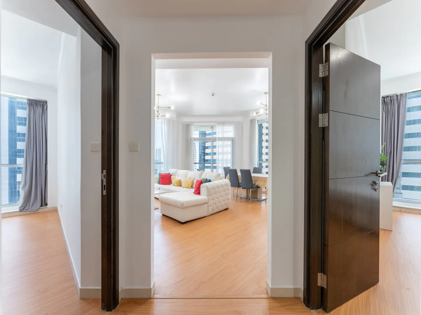 View through doorways into a modern apartment with wood floors, white walls, and city views. A white sofa with colorful pillows is visible.