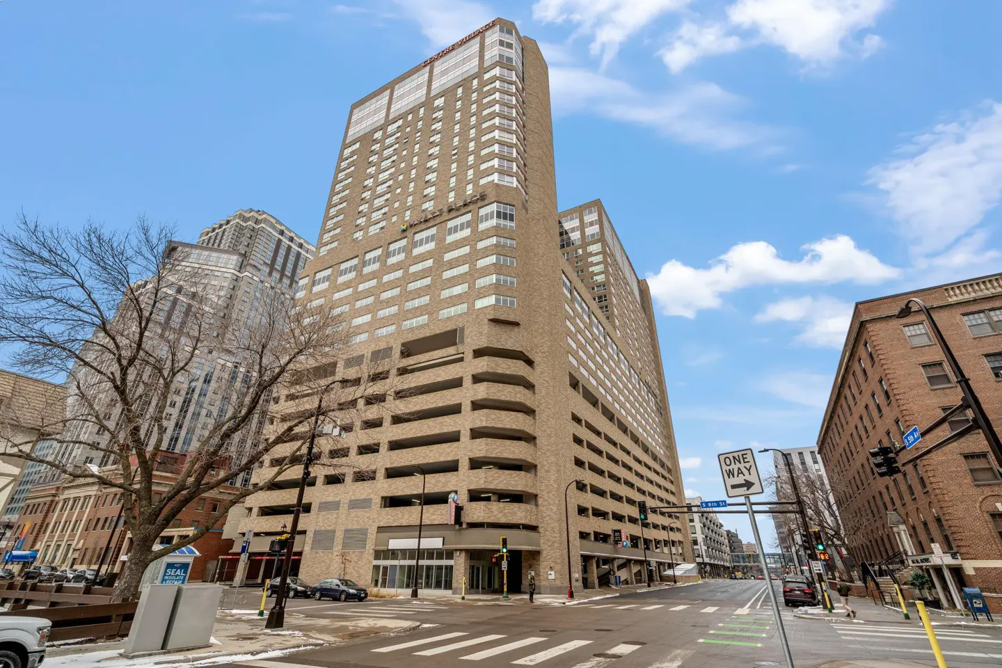 Cityscape view of a tall, tan brick building with a parking garage, street, and blue sky.