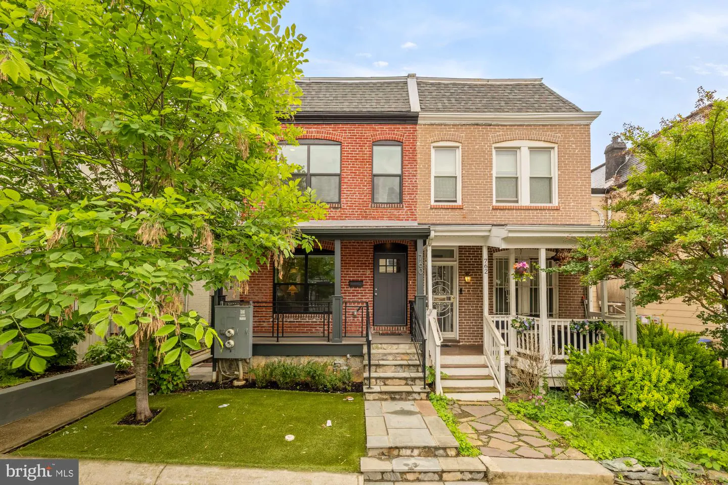Two-story brick row houses, one red and one tan, with front porches and steps leading up to the doors. A large green tree is on the left.