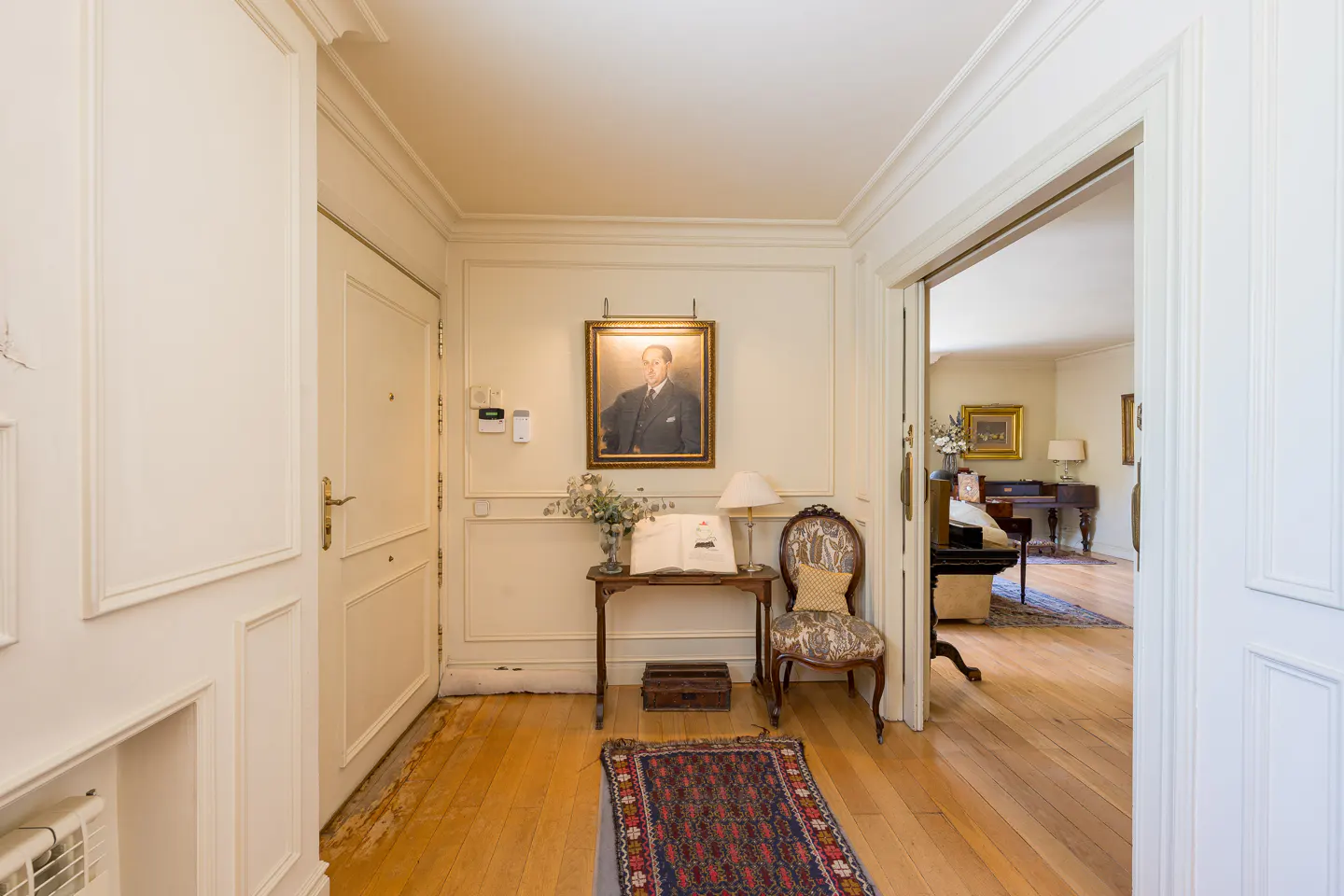 Entryway with white walls, wood floors, and a portrait above a table with a lamp and book. A patterned chair sits nearby. A doorway leads to a living room.