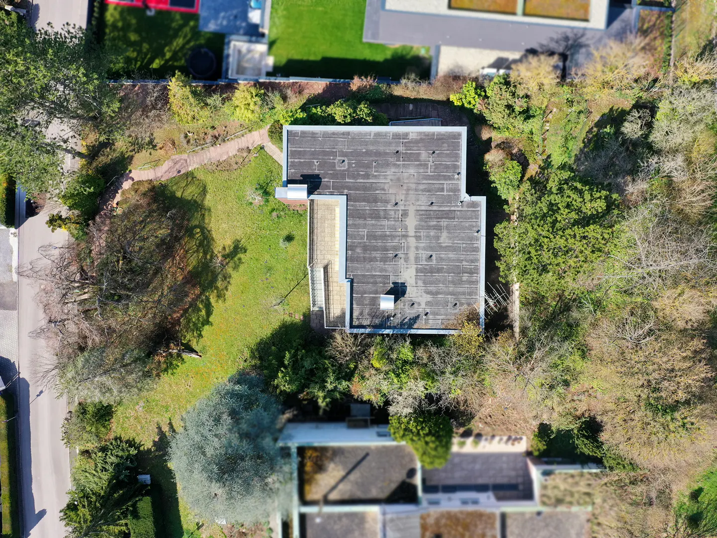 Aerial view of a modern, flat-roofed house with a dark gray roof, surrounded by green lawn and trees. A stone path leads to the house.