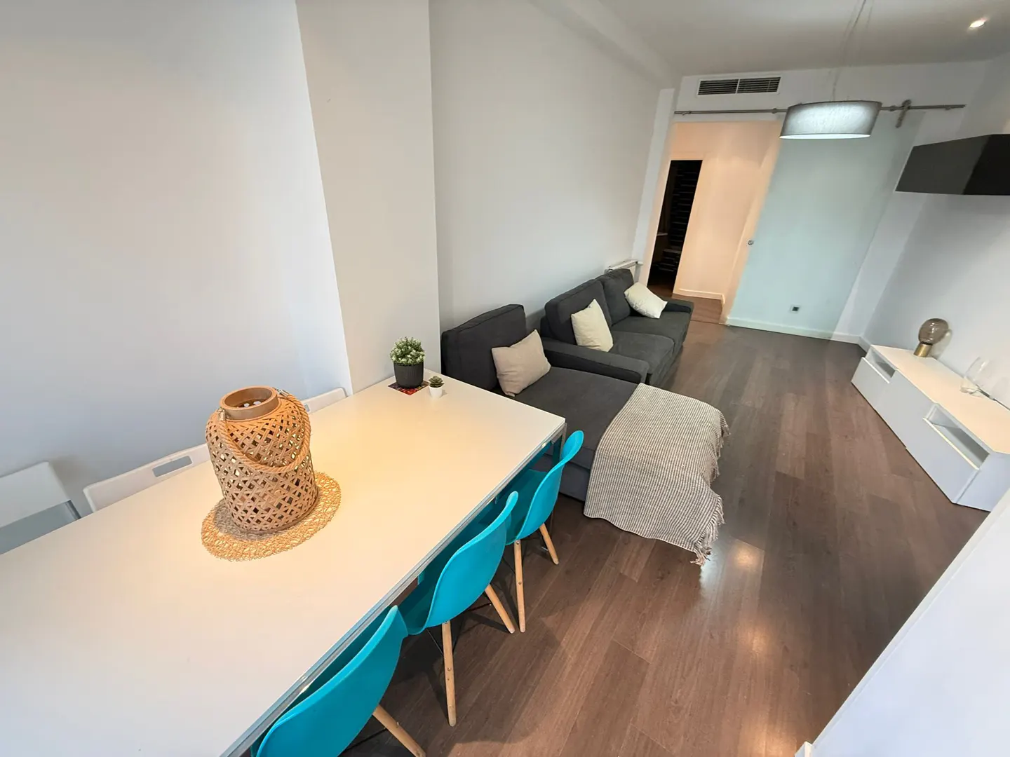 Bright living space with white walls, wood floors, and a gray sofa. A white table with blue chairs sits near a woven lantern.