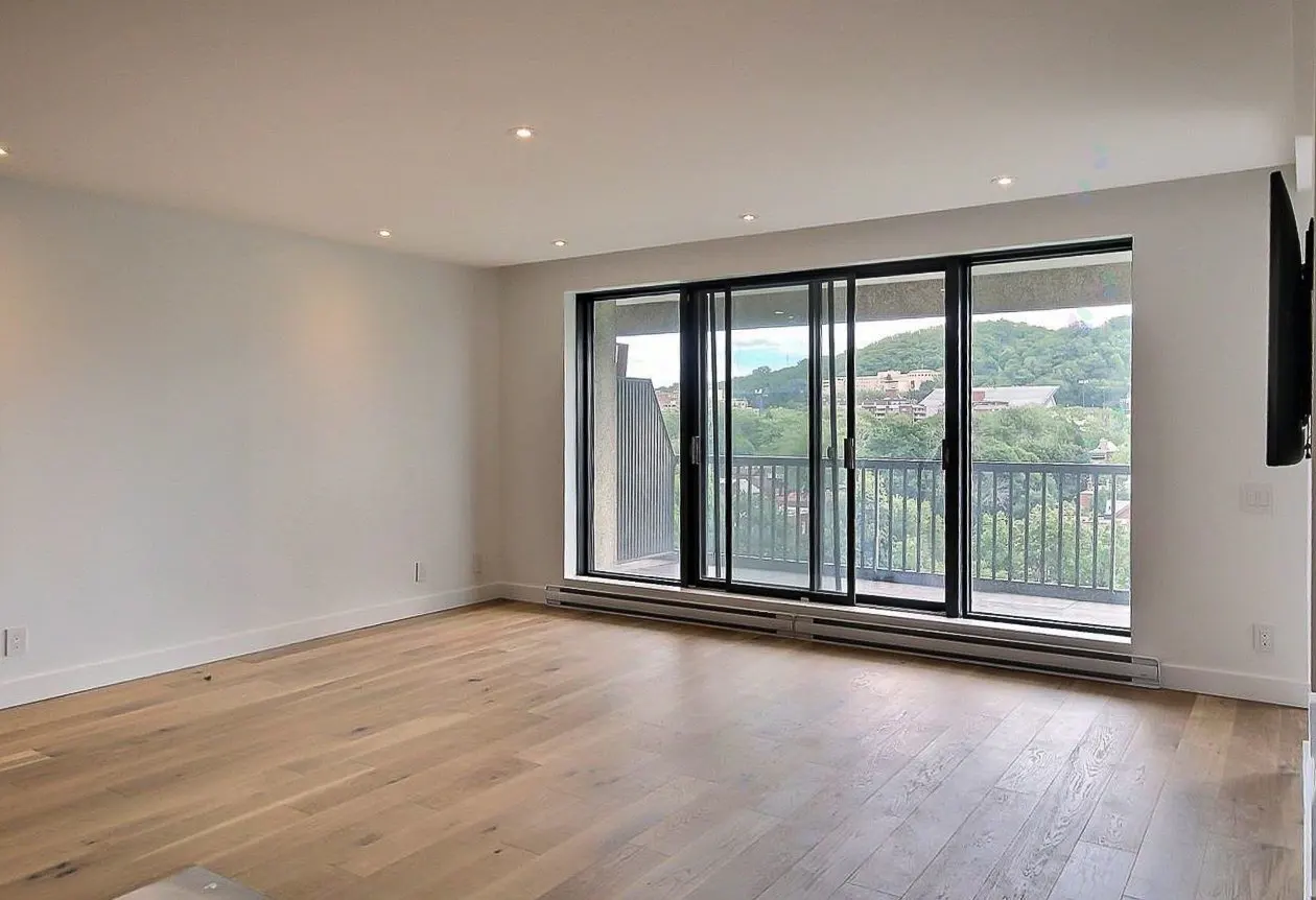 Bright, empty room with light wood floors and white walls. Large sliding glass doors lead to a balcony with a view of trees and hills.