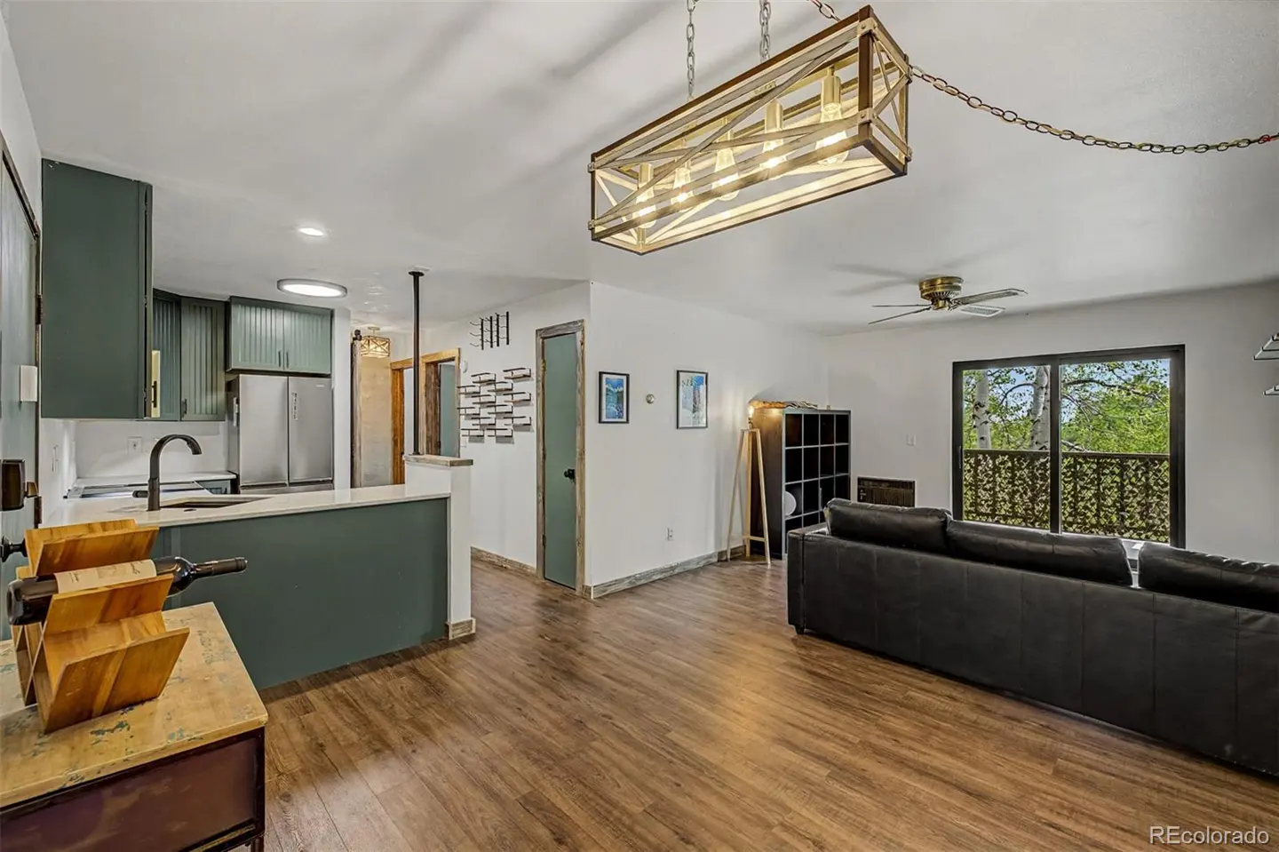 Open-concept living space with wood floors, a black leather sofa, and a kitchen with green cabinets. A rectangular chandelier hangs above.