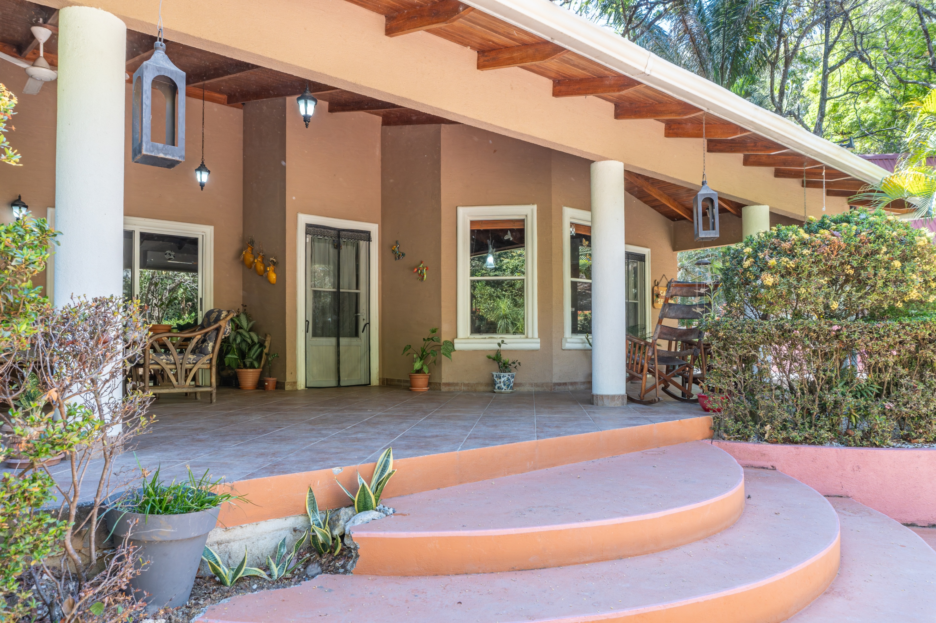 Covered porch of a tan house with white columns, hanging lanterns, and a wooden rocking chair. Three steps lead up to the porch.