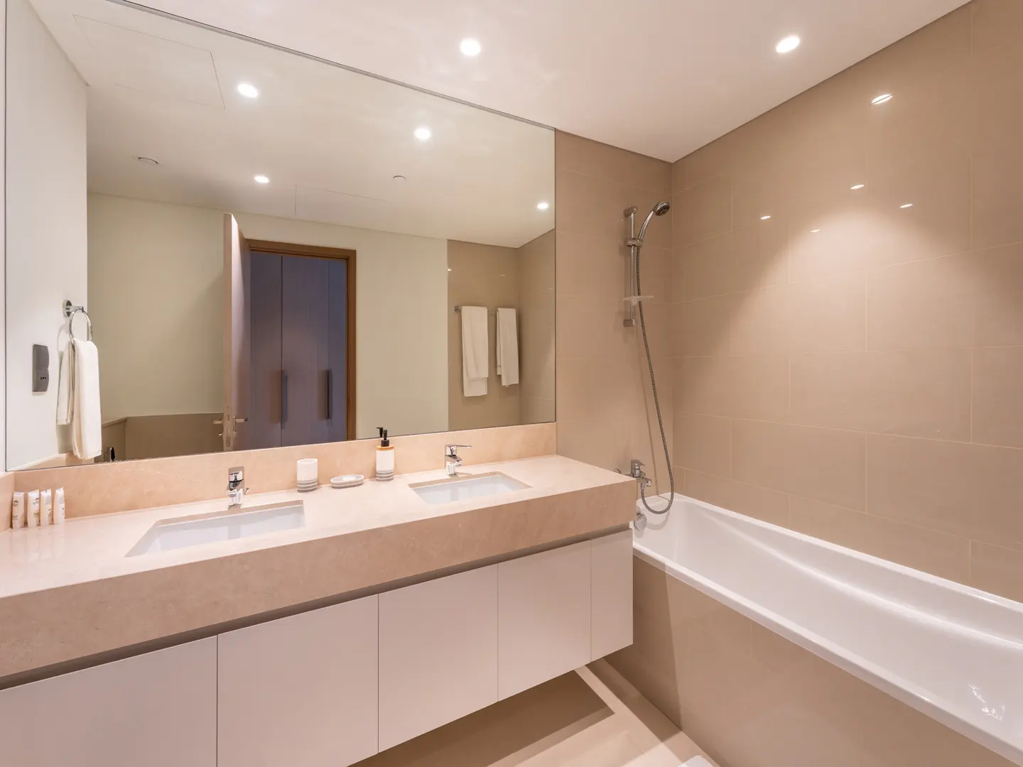 Modern bathroom with beige tile walls, white cabinets, and a double sink vanity with a large mirror above. A white bathtub is on the right.