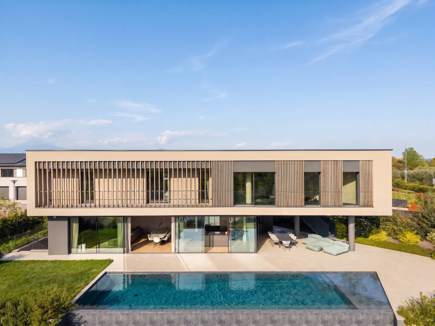 Modern beige house with a blue pool, green lawn, and patio furniture under a blue sky.