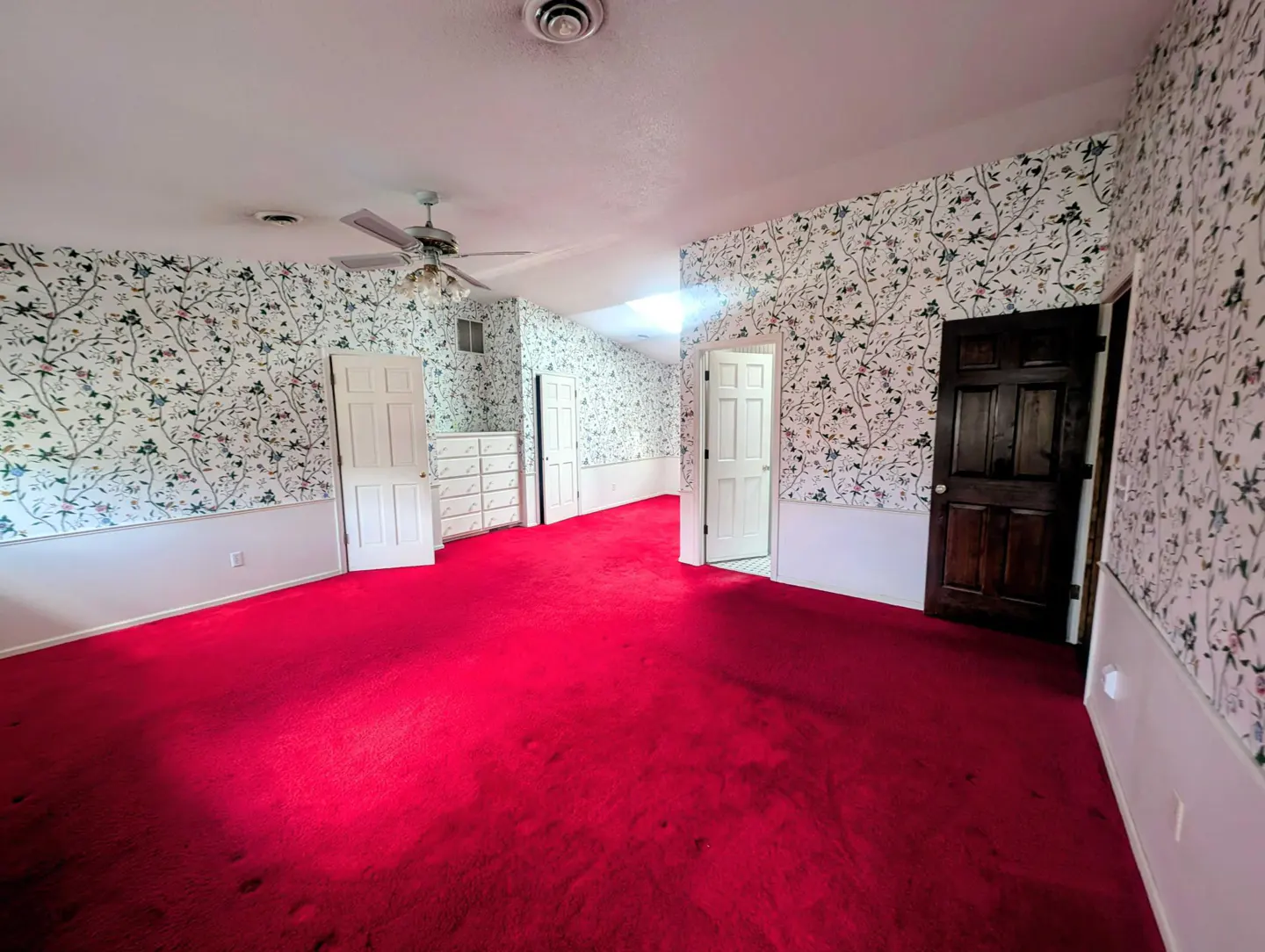 A bedroom with red carpet, floral wallpaper, white trim, and a dark wood door.